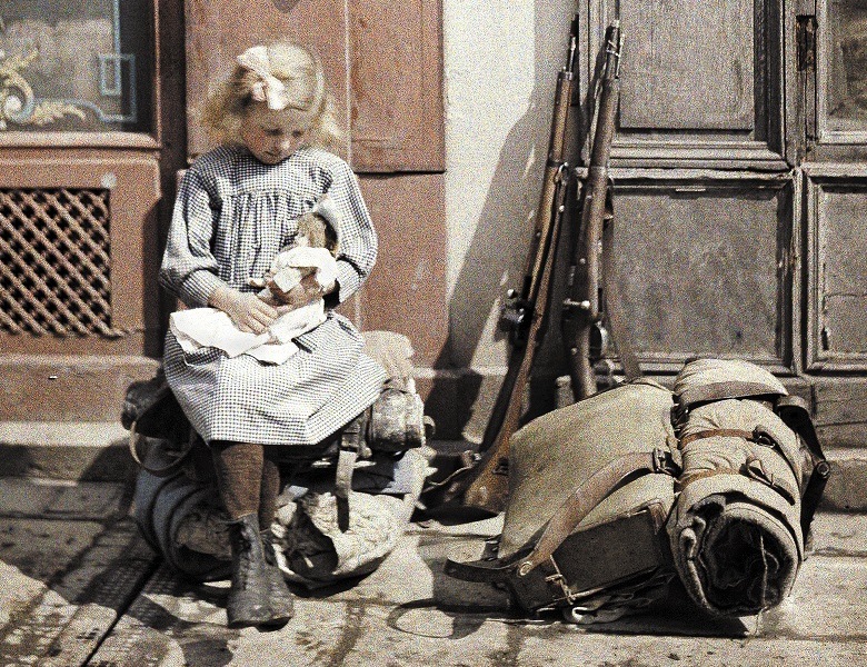A little girl playing with her doll; two guns and a knapsack are next to her on the ground, 1917 (autochrome) by Fernand Cuville